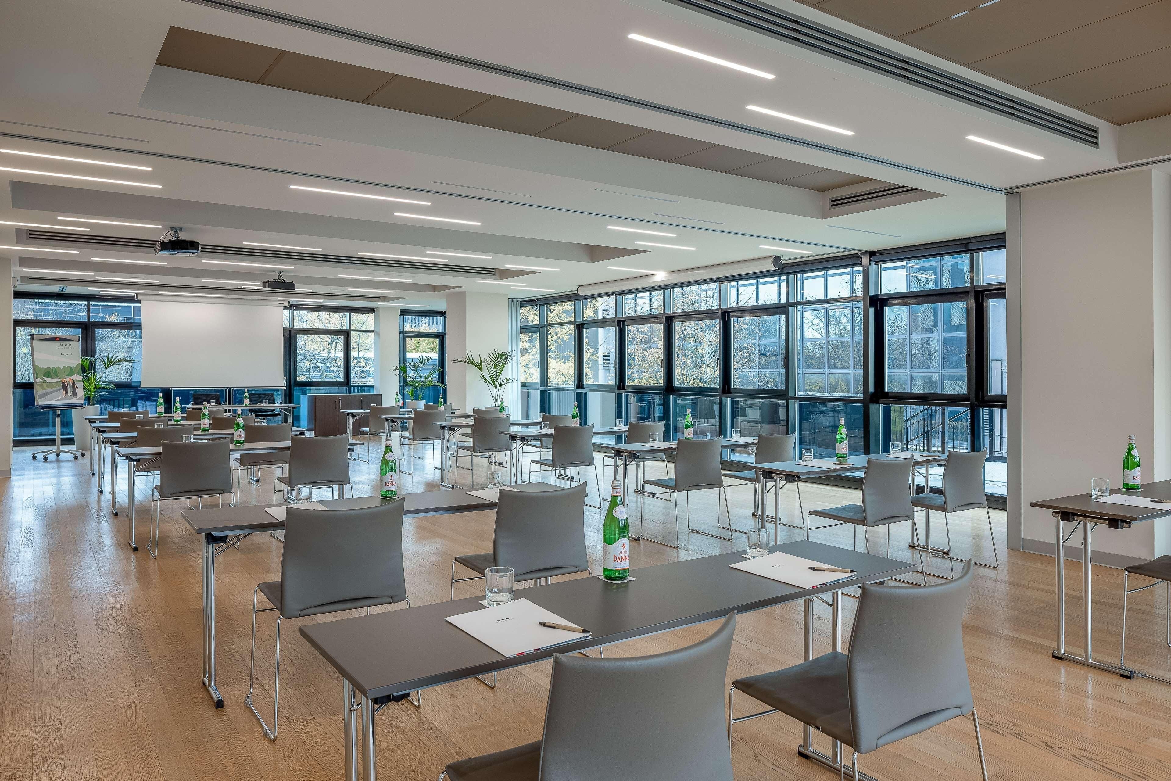 A spacious conference room with multiple tables and chairs, large windows, and ceiling-mounted lighting.