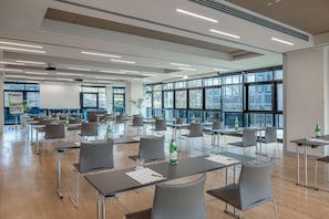A spacious conference room with multiple tables and chairs, large windows, and ceiling-mounted lighting. - UNA Hotels Bologna Fiera (Bologna)