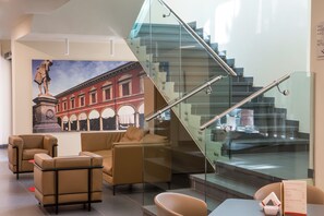 A modern interior with a glass staircase, leather sofas, and a mural of a historical building. - UNA Hotels Bologna Fiera (Bologna)