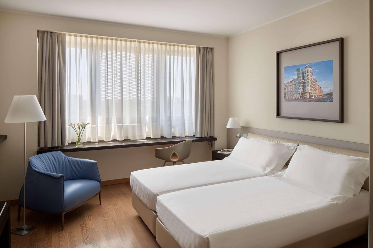 Hotellihuone, jossa on kaksi sänkyä, työpöytä, tuoli ja ikkuna, jossa on verhot.