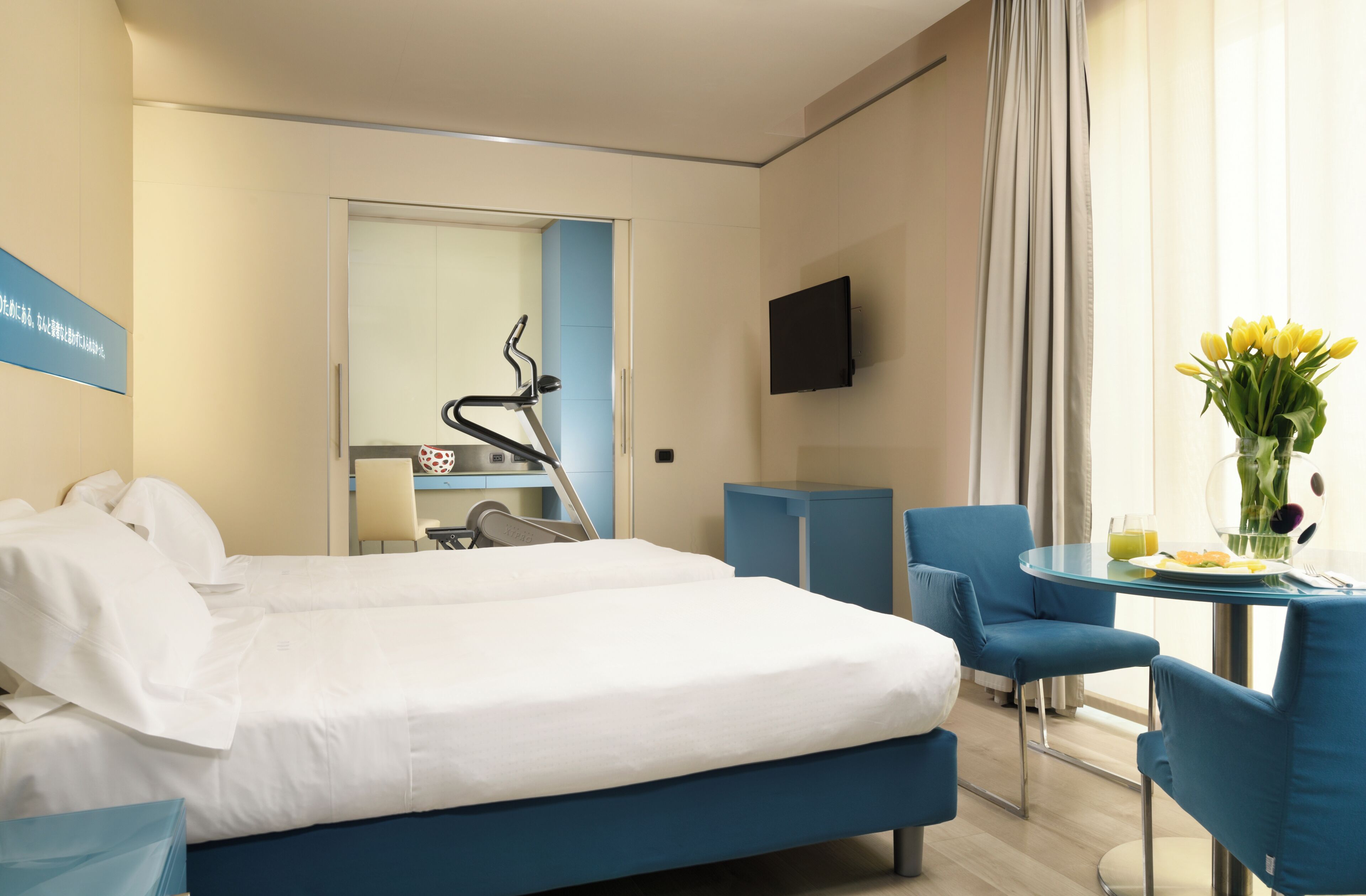 Habitación de hotel con cama, escritorio y silla, televisor, mesa de comedor con sillas y ventana con cortinas.