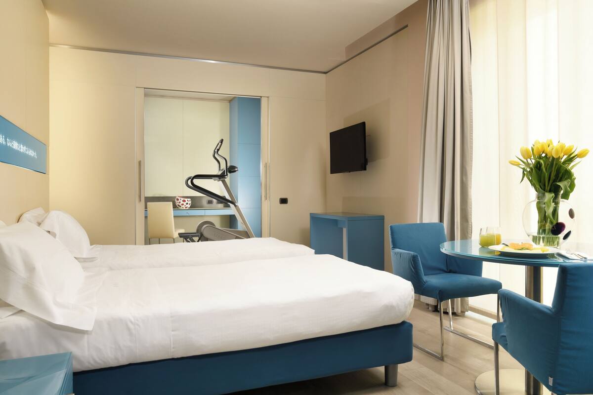 Habitación de hotel con cama, escritorio y silla, televisor, mesa de comedor con sillas y ventana con cortinas.