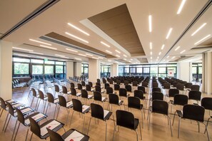 Uma sala de conferências ampla, com fileiras de cadeiras e mesas, janelas grandes e luminárias modernas.