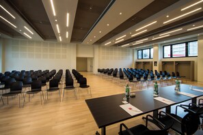 Un salón de conferencias con filas de sillas, una mesa larga con documentos y una pantalla de proyector.