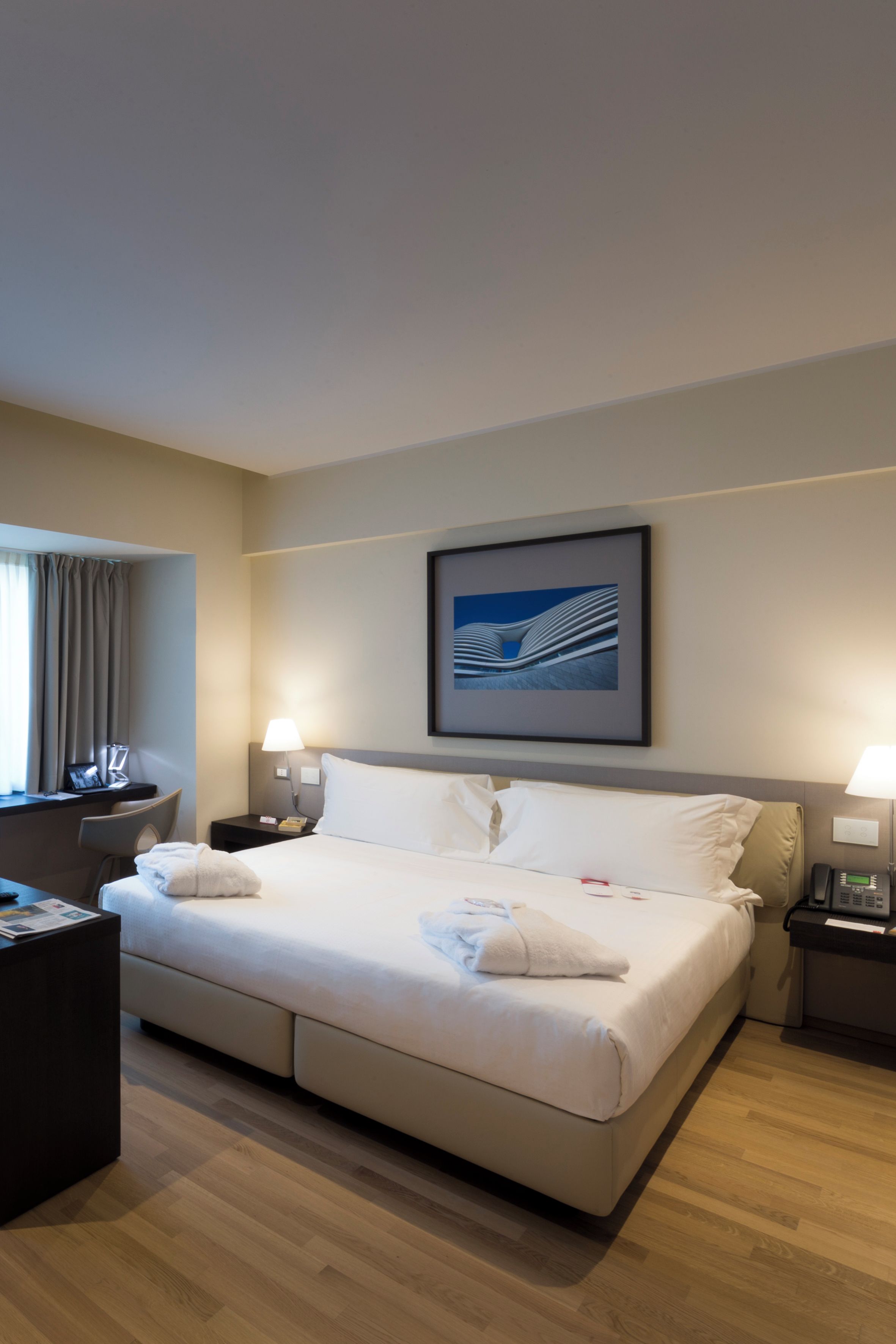 A hotel room with a bed, bedside tables, a desk, a chair, a window with curtains, and a framed picture on the wall.