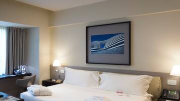 A hotel room with a bed, bedside tables, a desk, a chair, a window with curtains, and a framed picture on the wall.