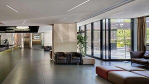Un vestíbulo amplio con diseño moderno, que incluye un mostrador de recepción, un área de descanso con sofás y otomanas, ventanas grandes con vistas al exterior y una zona de bar al fondo.