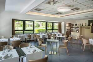 Un restaurant doté de nombreuses tables et chaises, de grandes fenêtres offrant une vue sur la verdure et d’un présentoir à bouteilles.