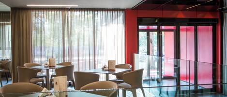 A modern dining area with tables and chairs, large windows with sheer curtains, and a red accent wall.