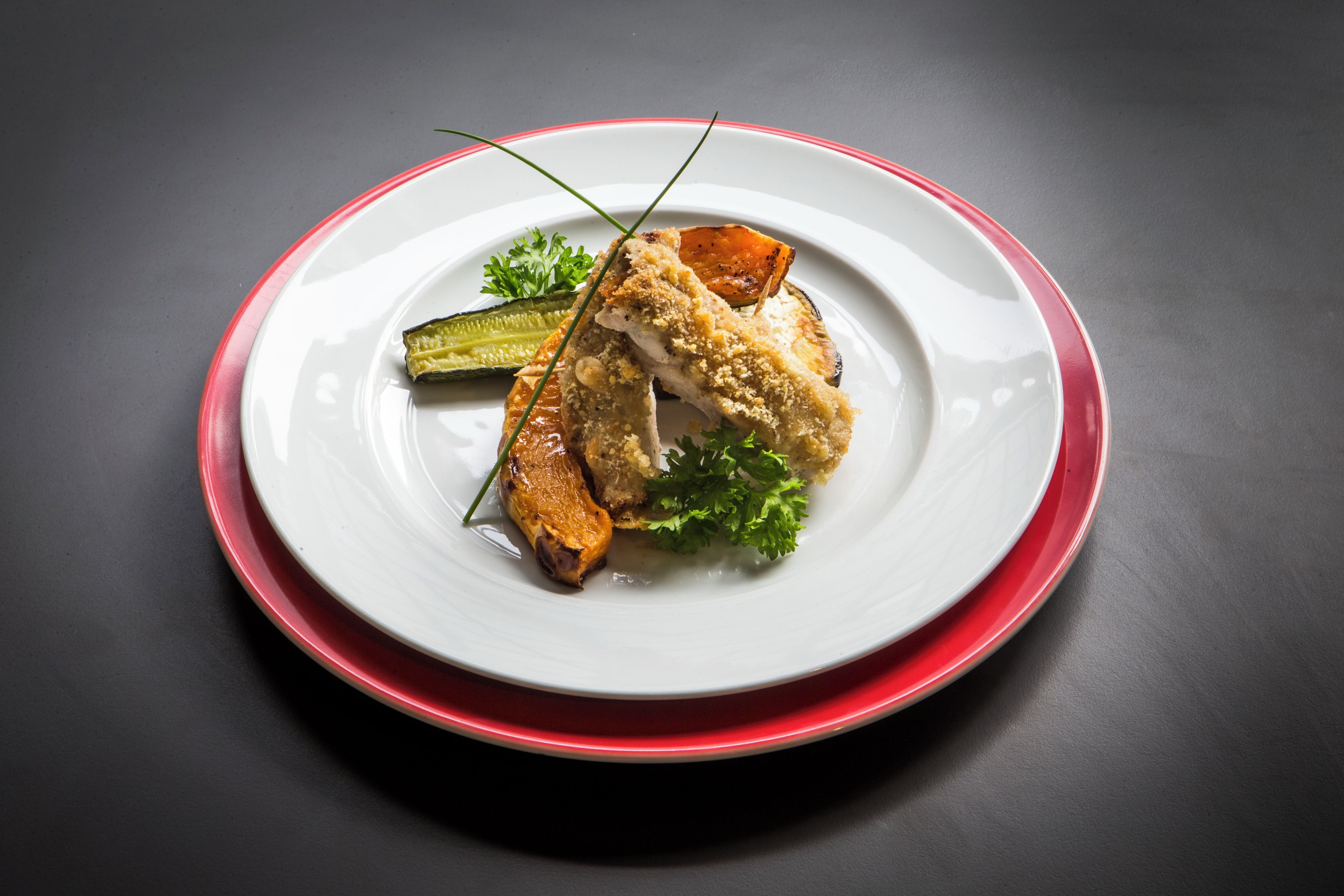 A plate with a piece of breaded chicken, a slice of tomato, and a garnish on a white plate with a red rim, set against a dark background.
