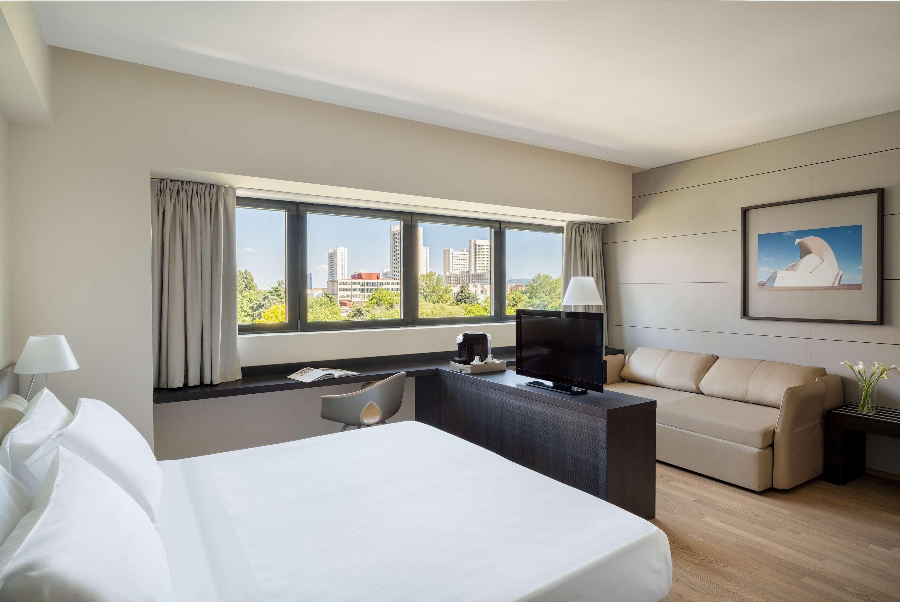 Una camera d'albergo con un letto, una scrivania, una televisione, un divano e una finestra con vista sulla città.