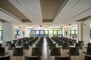 Une salle de conférence avec des rangées de chaises, de grandes fenêtres et des bouches d’aération au plafond.
