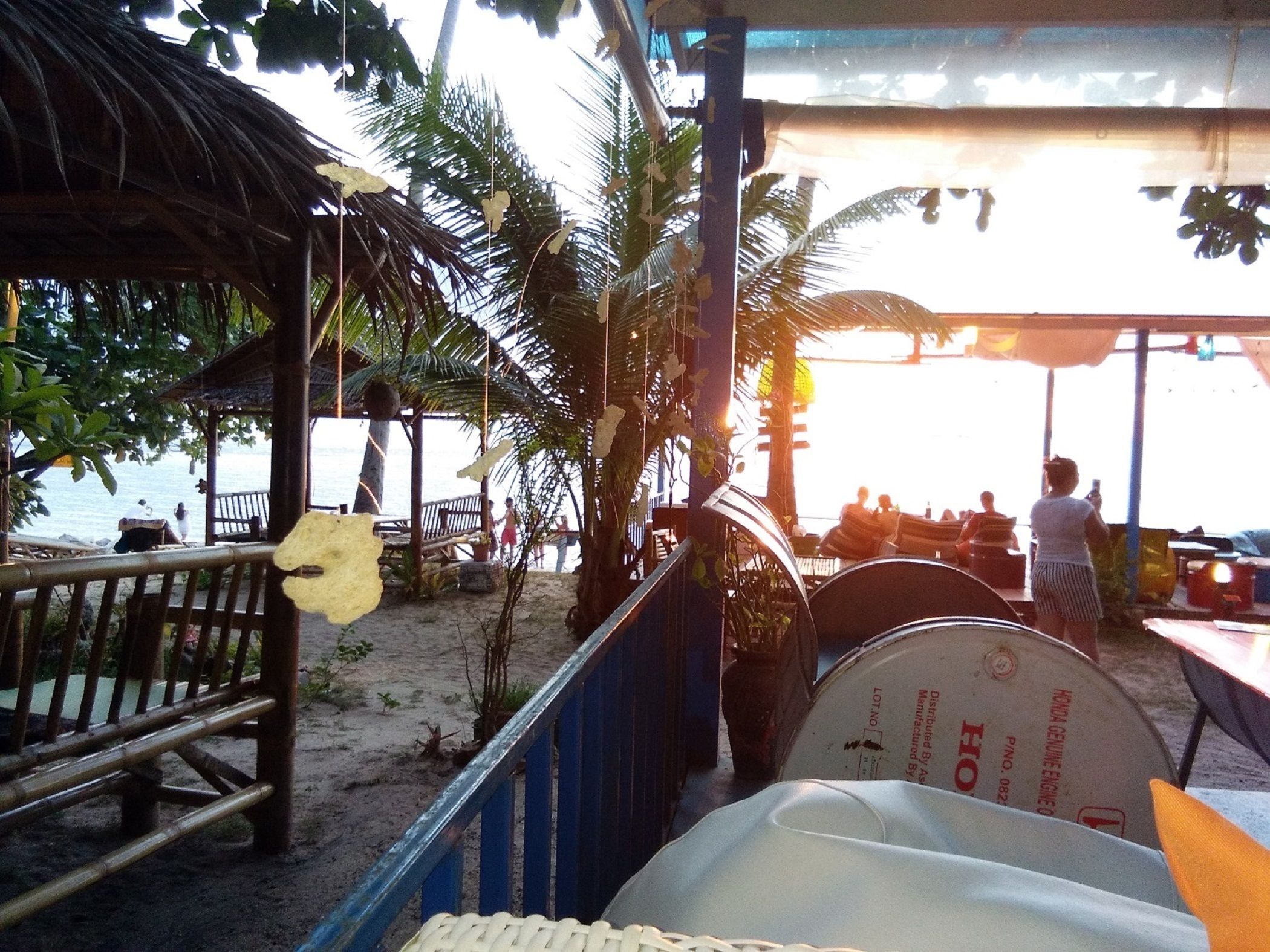 A beachside restaurant with outdoor seating, a thatched roof structure, and palm trees.