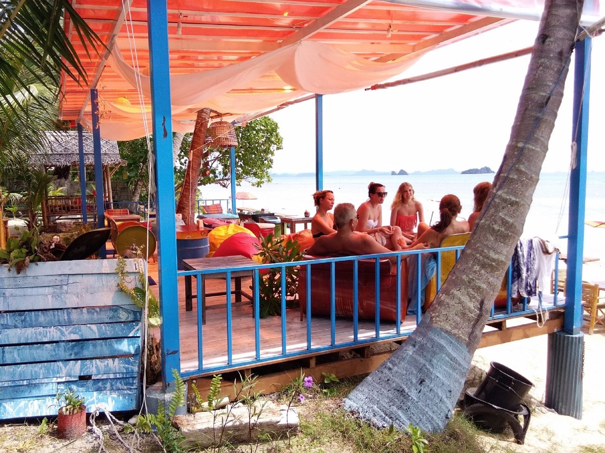 A beachside seating area with a view of the sea, covered by a canopy, and surrounded by palm trees.