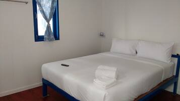 A single-bed room with a wooden frame, white bedding, a remote control, and a window with blue curtains.
