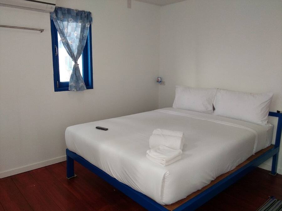 A single-bed room with a wooden frame, white bedding, a remote control, and a window with blue curtains.