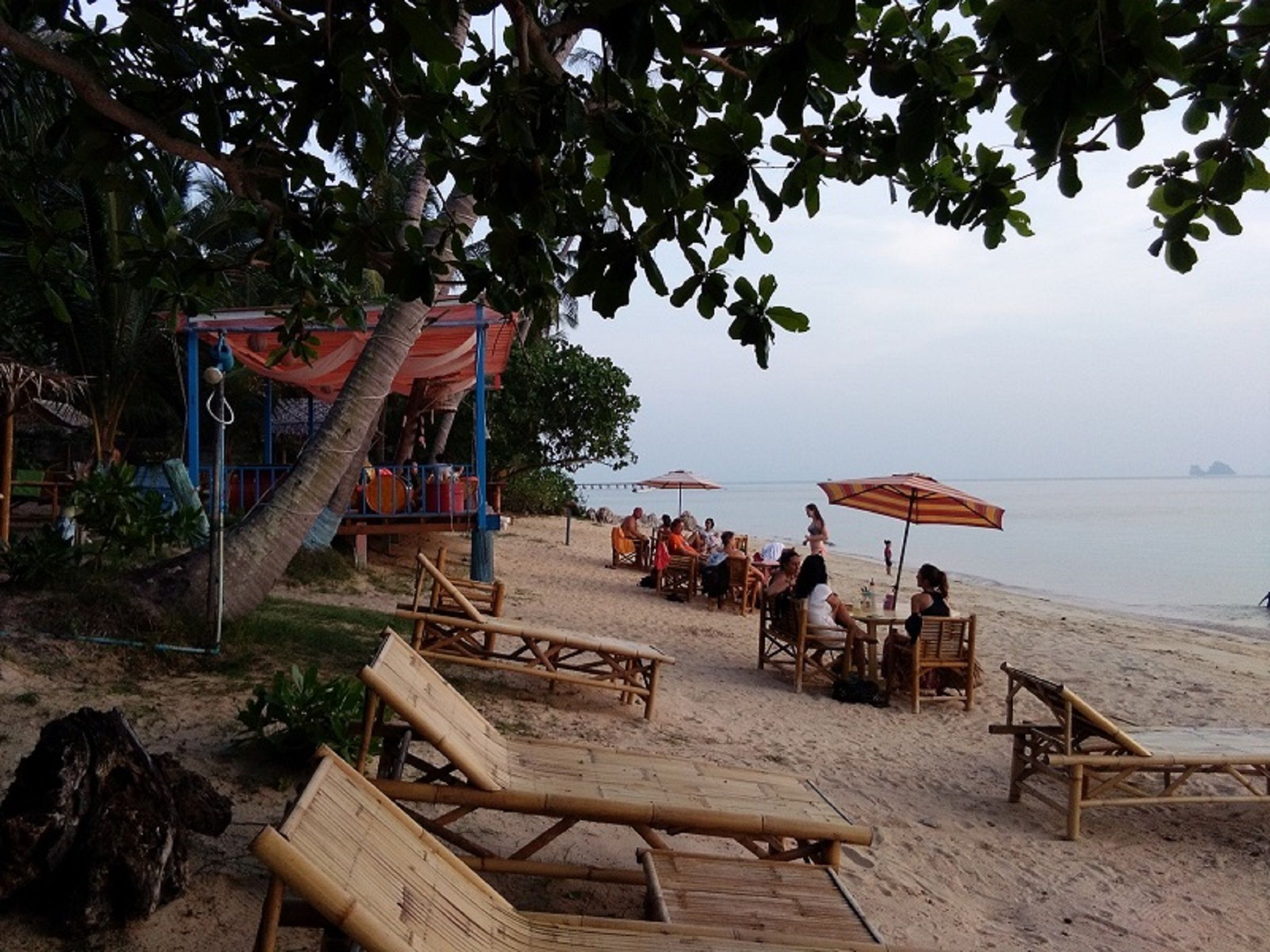 A beach with bamboo lounge chairs and umbrellas, a shaded area with a red canopy, and a view of the sea.