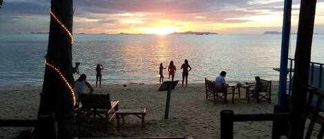 A beachside seating area with wooden benches and tables, overlooking the sea during sunset.