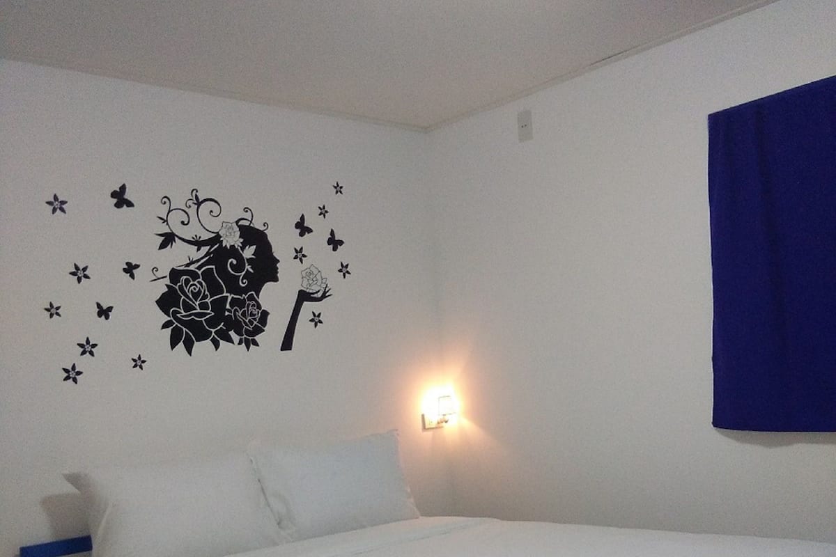 A single bed with white bedding and a blue frame, a wall decal with a silhouette of a woman holding a rose, and a wall-mounted light.