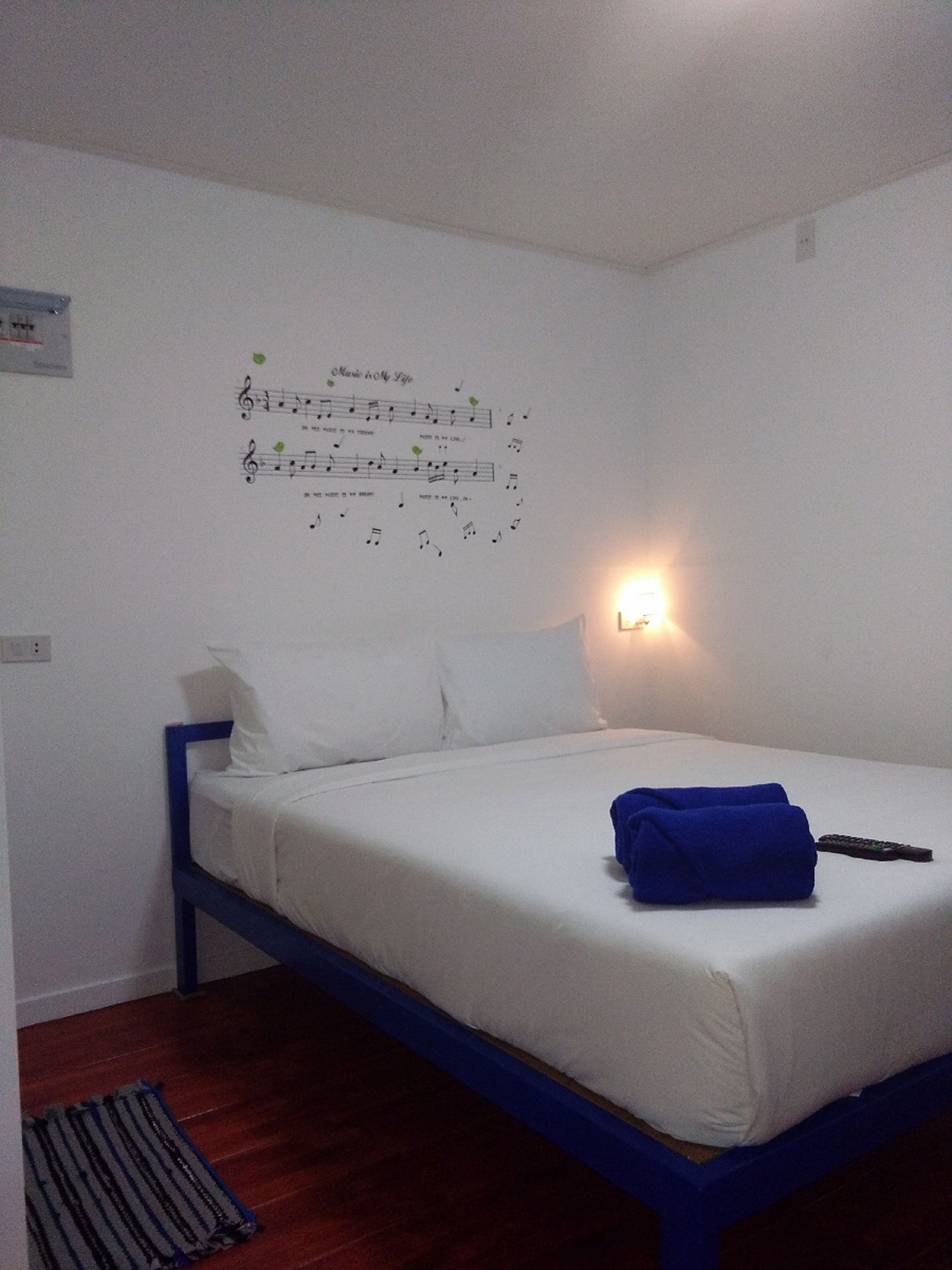 A single bed with white bedding and a blue frame, a wall-mounted light, and a poster with musical notes.