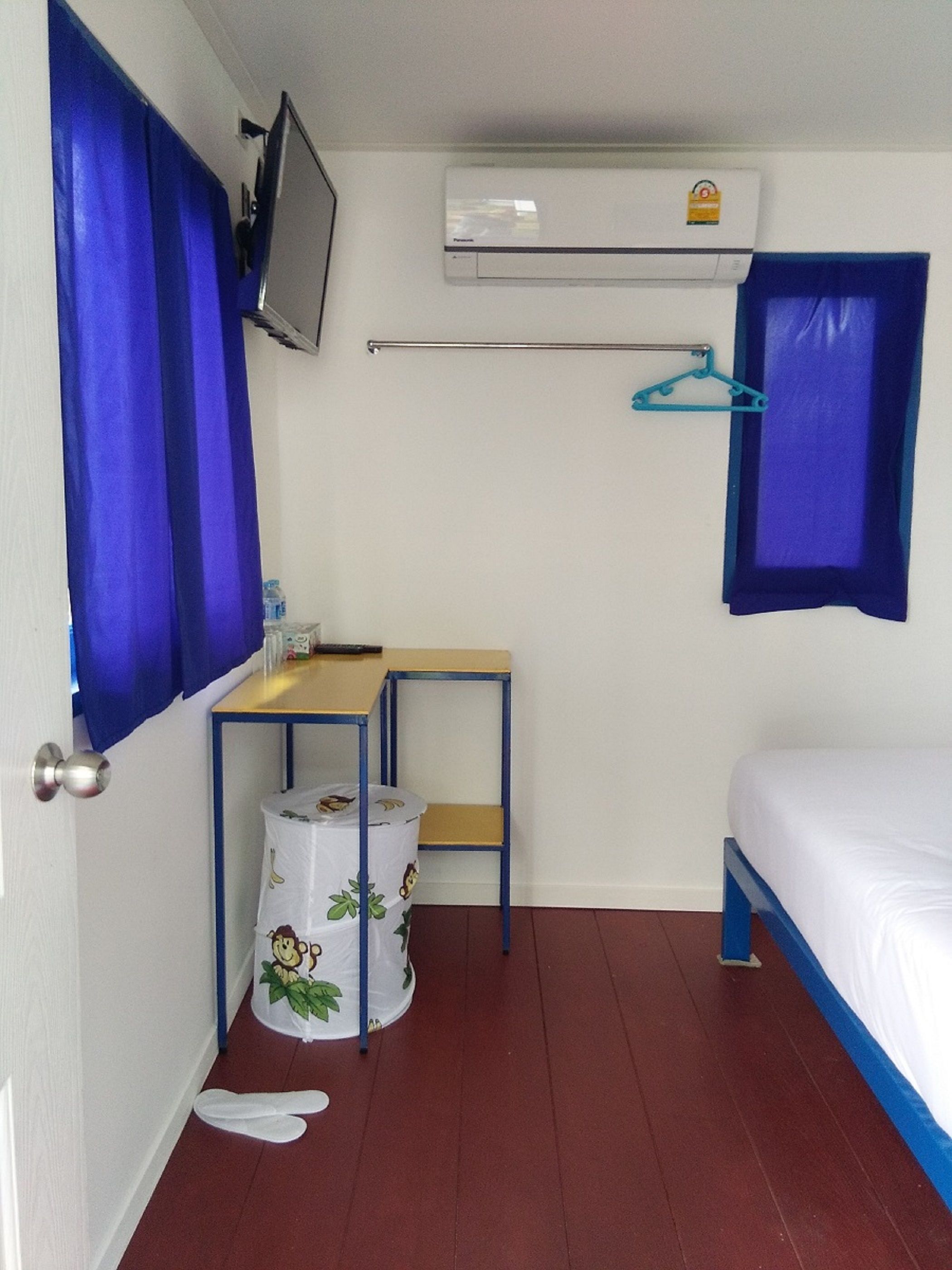 Habitació petita i senzilla amb llit, escriptori, cadira, televisió penjada a la paret i finestra amb cortines blaves.