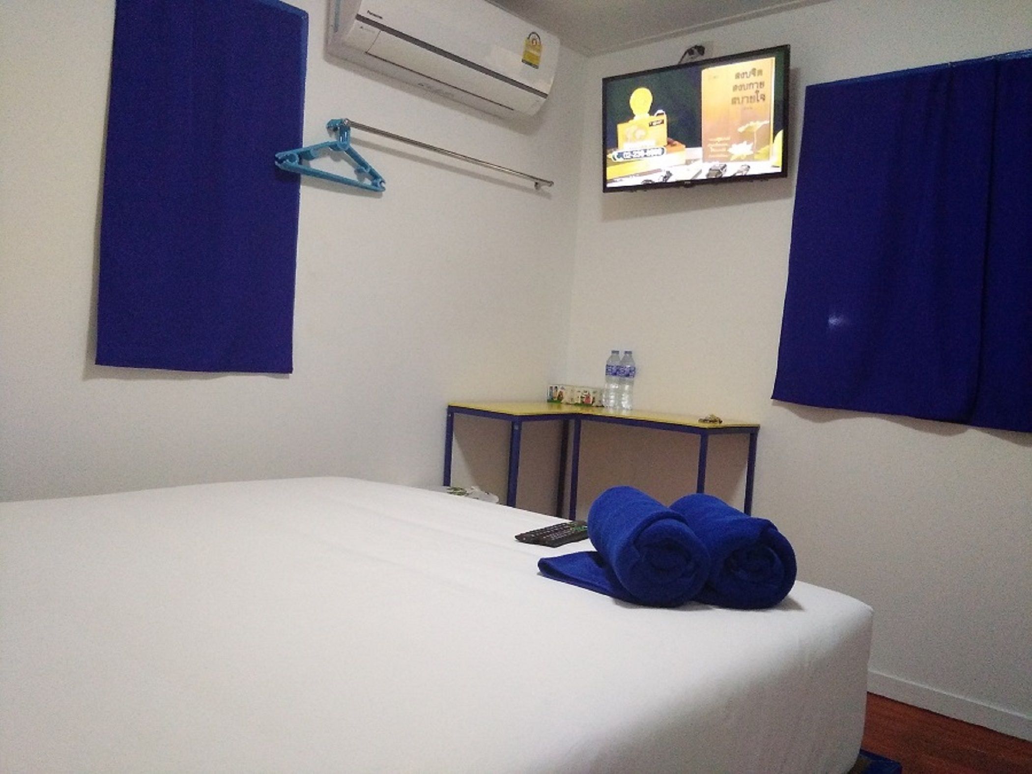 A hotel room with a bed, a TV mounted on the wall, a small table with a water bottle, and a blue towel on the bed.