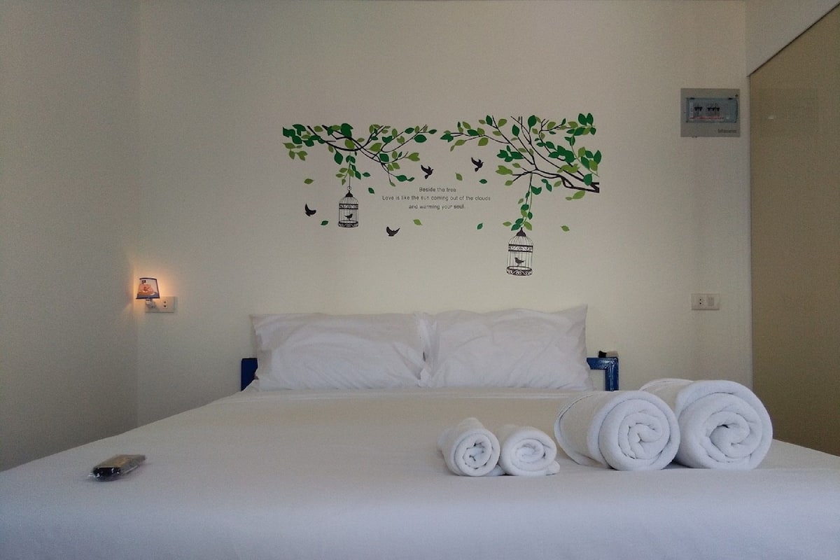A neatly made bed with white linens and rolled towels, a wall-mounted light fixture, and a decorative wall art featuring birds and a tree.