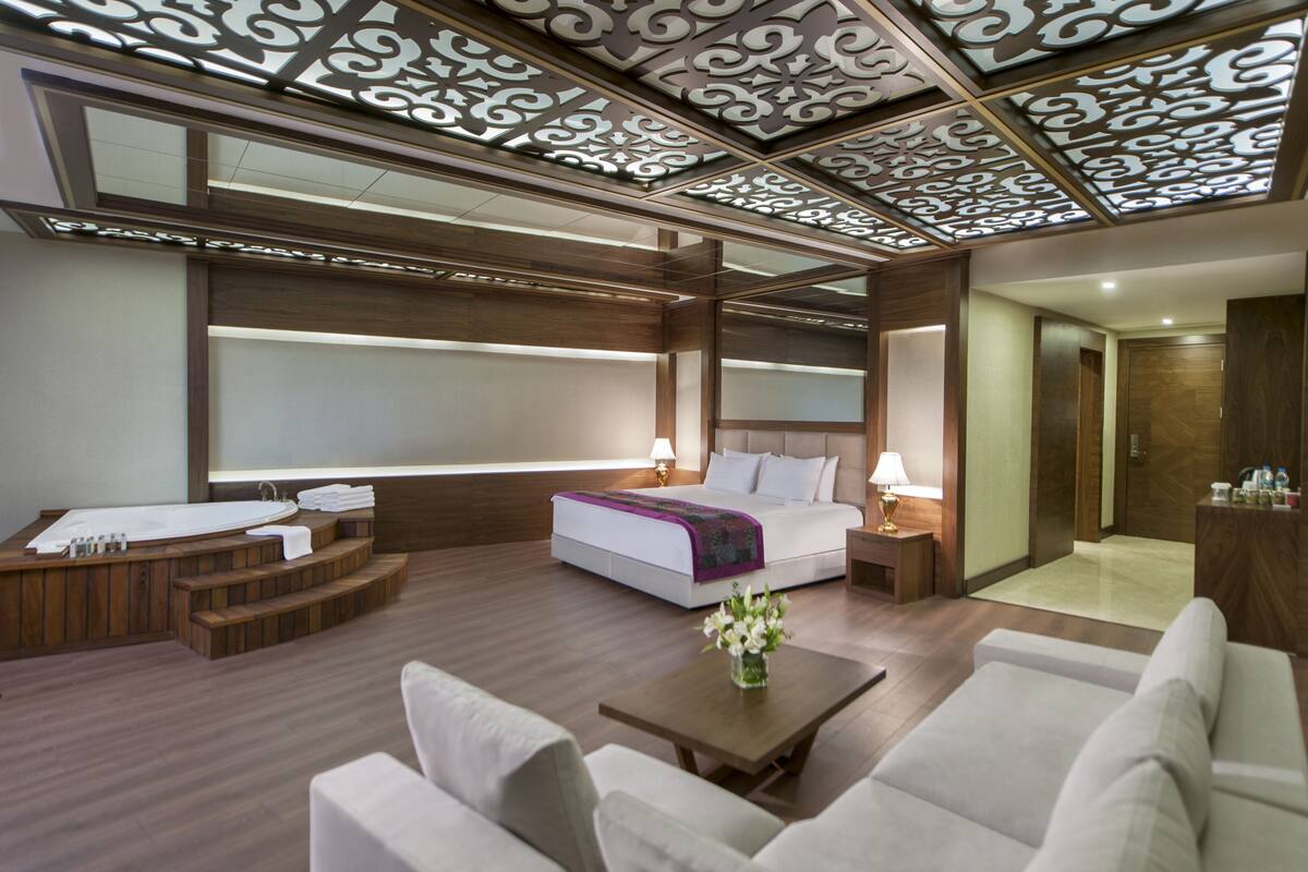 A modern hotel room with a large bed, a sitting area with a coffee table, a small bathroom with a bathtub, and a decorative ceiling.
