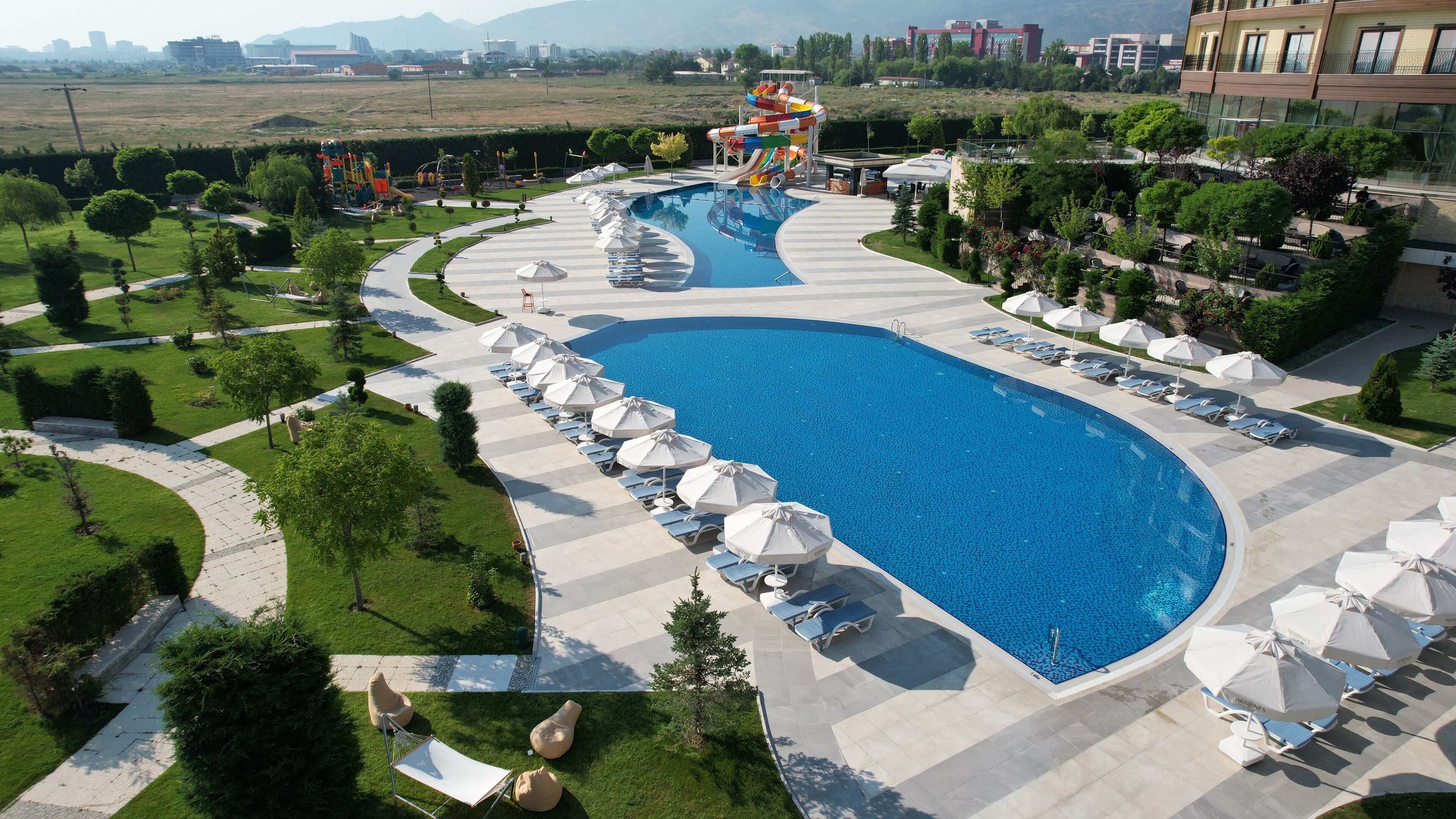 A large swimming pool with a water slide, surrounded by lounge chairs and umbrellas, with a landscaped garden and distant buildings.
