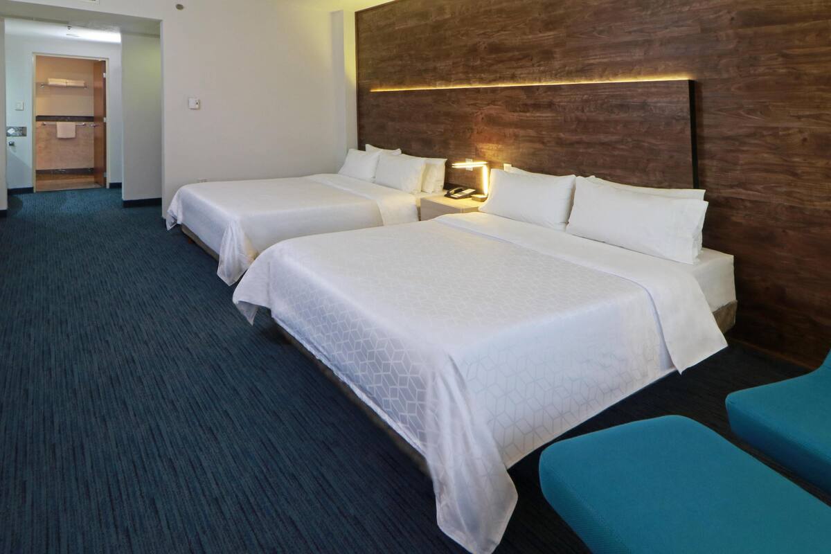 A hotel room with two beds, a wooden headboard, a nightstand with a lamp, and a blue chair.