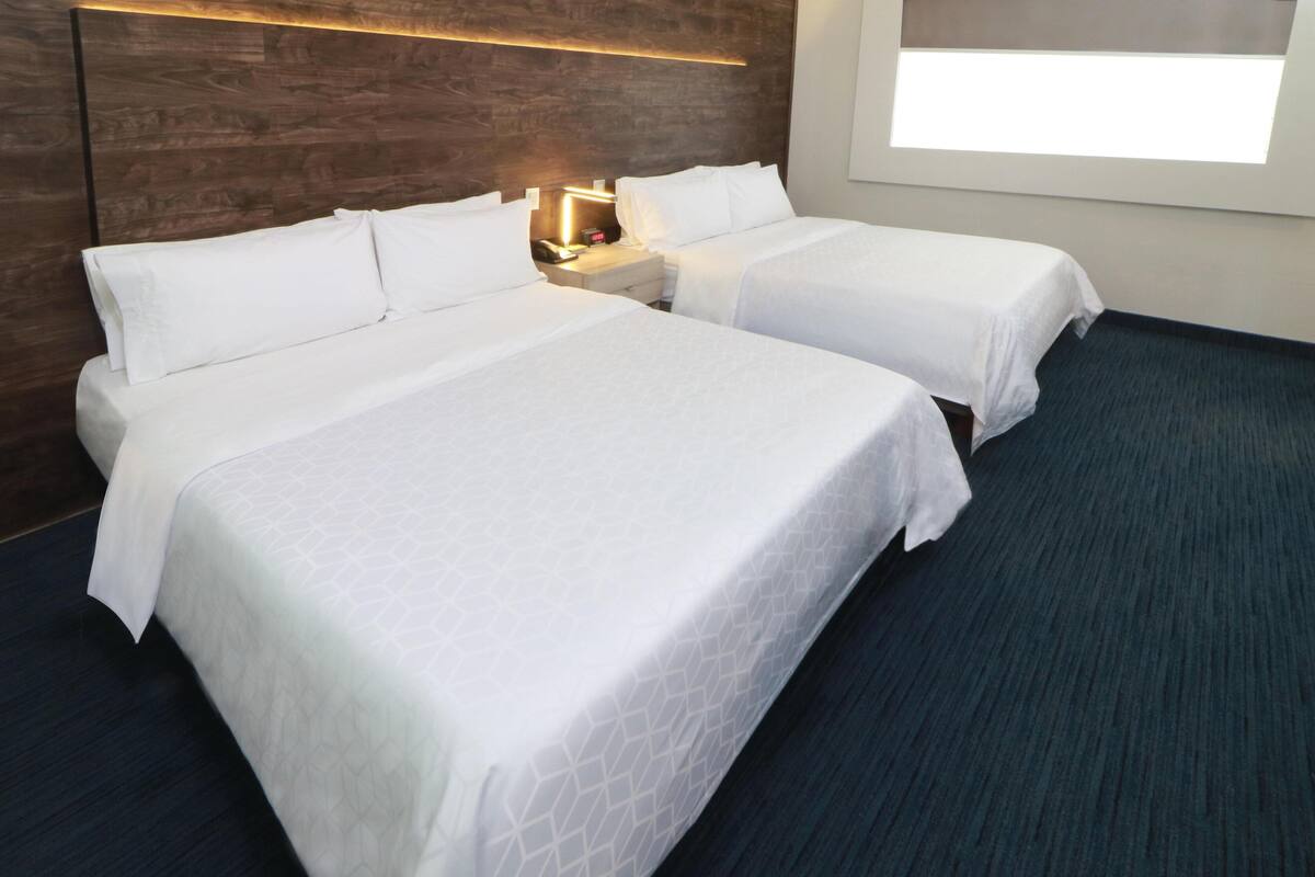 A hotel room with two beds, a wooden headboard, a nightstand with a lamp, and a window.