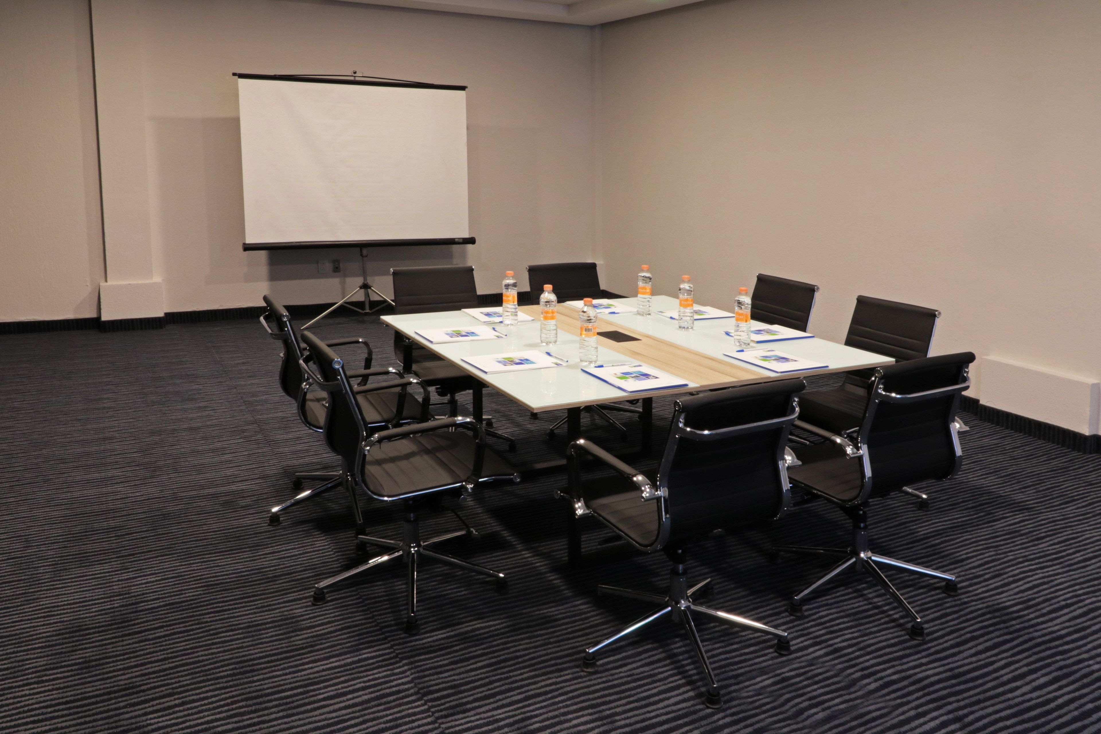 A conference room with a long table, black chairs, a projector screen, and water bottles.