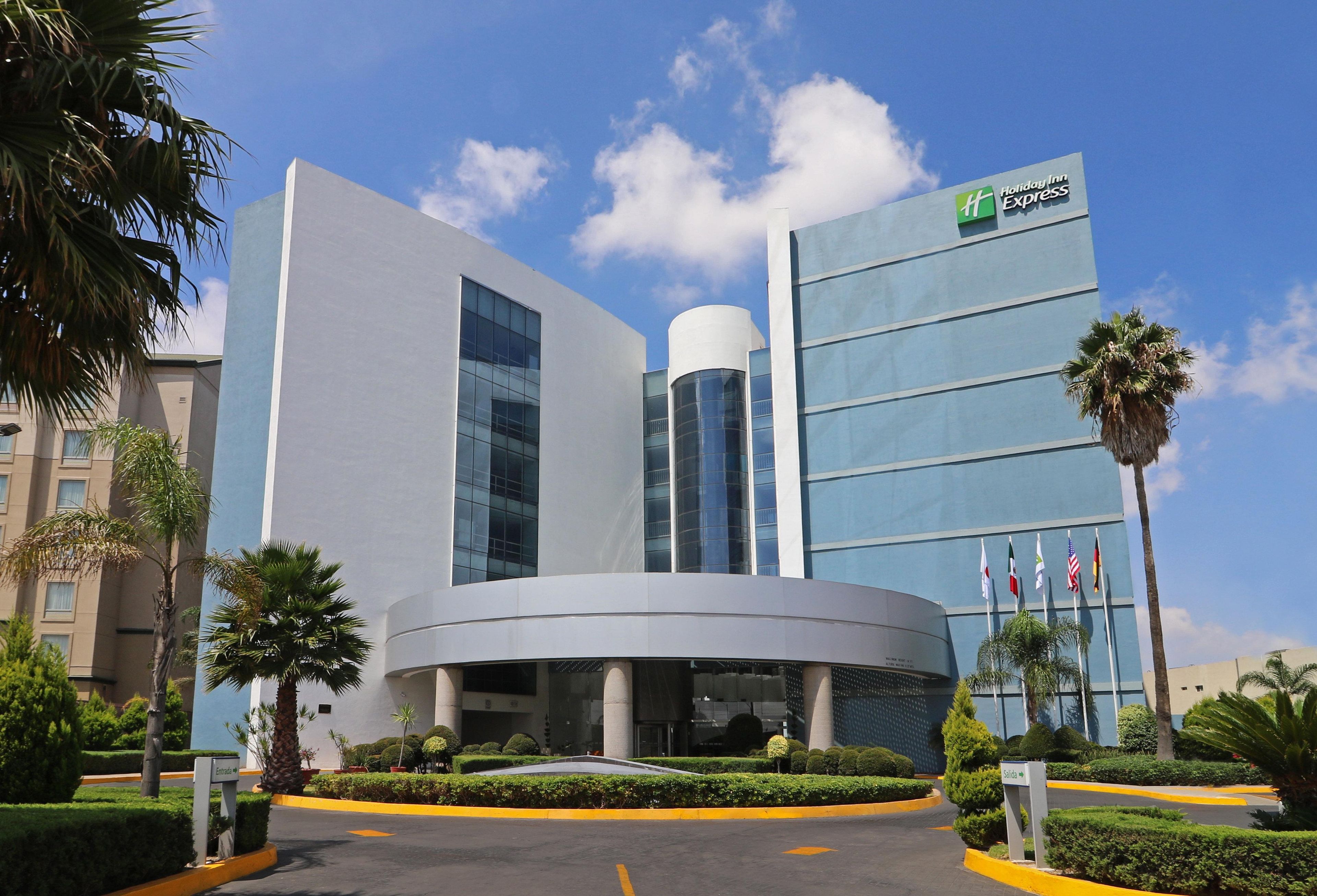 A modern hotel with a prominent Holiday Inn Express sign, a circular entrance, and a well-maintained garden with palm trees.