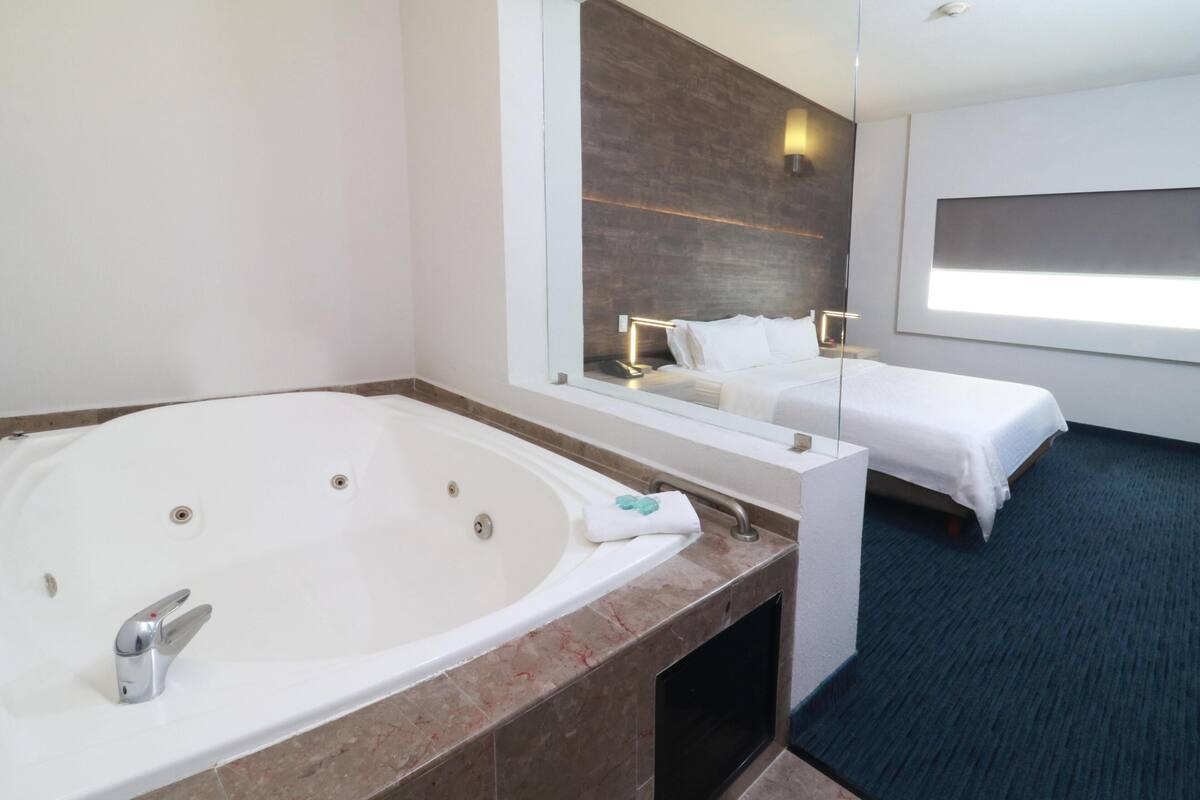 A modern hotel room with a large bathtub, a bed, and a large mirror reflecting the room.