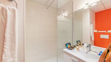 Un baño moderno con cabina de ducha de vidrio, un lavabo blanco con artículos de aseo y un espejo grande.
