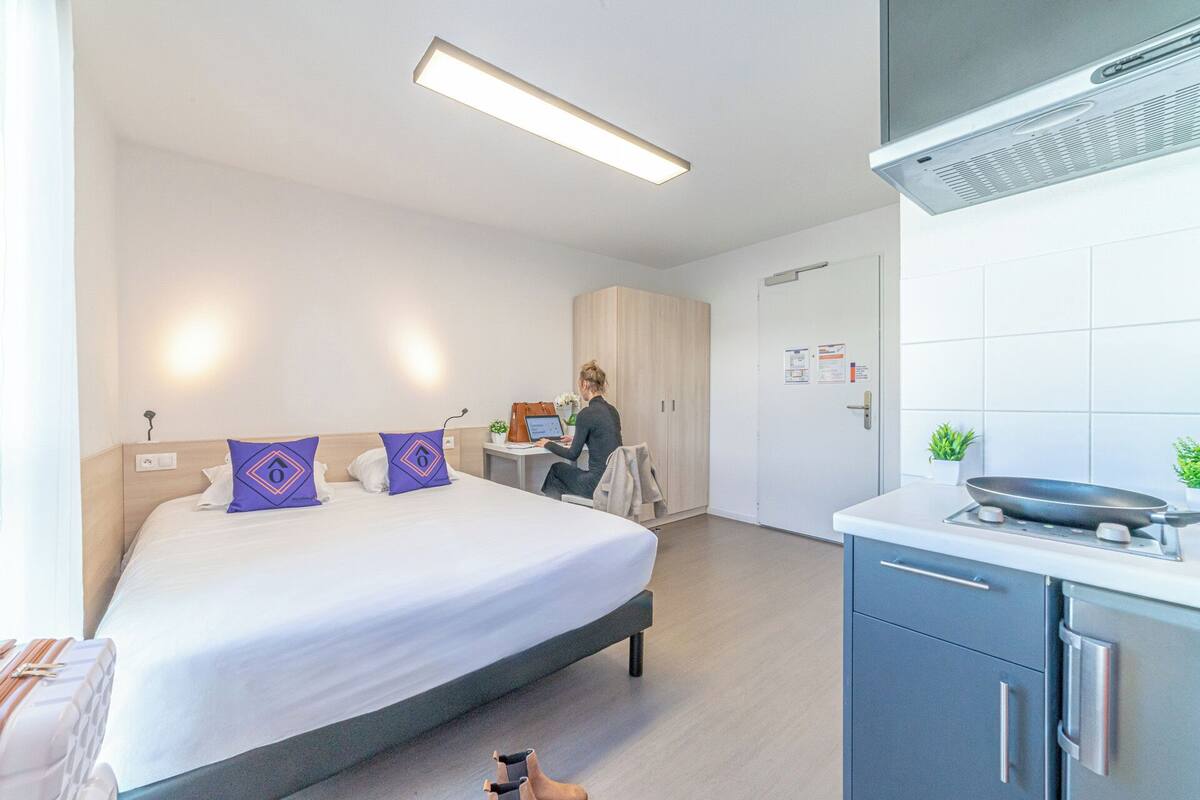 A modern hotel room with a bed, a desk, a kitchenette, and a bathroom.