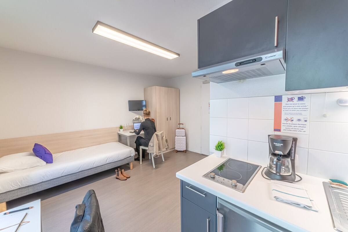 A compact hotel room with a bed, a desk, a kitchenette, and a TV.