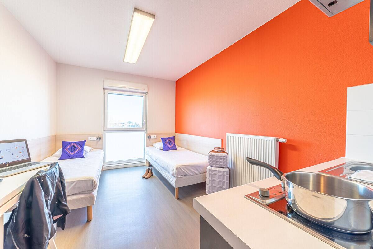 A hotel room with two single beds, a desk with a laptop, a suitcase, and a kitchenette with a pot on the stove.