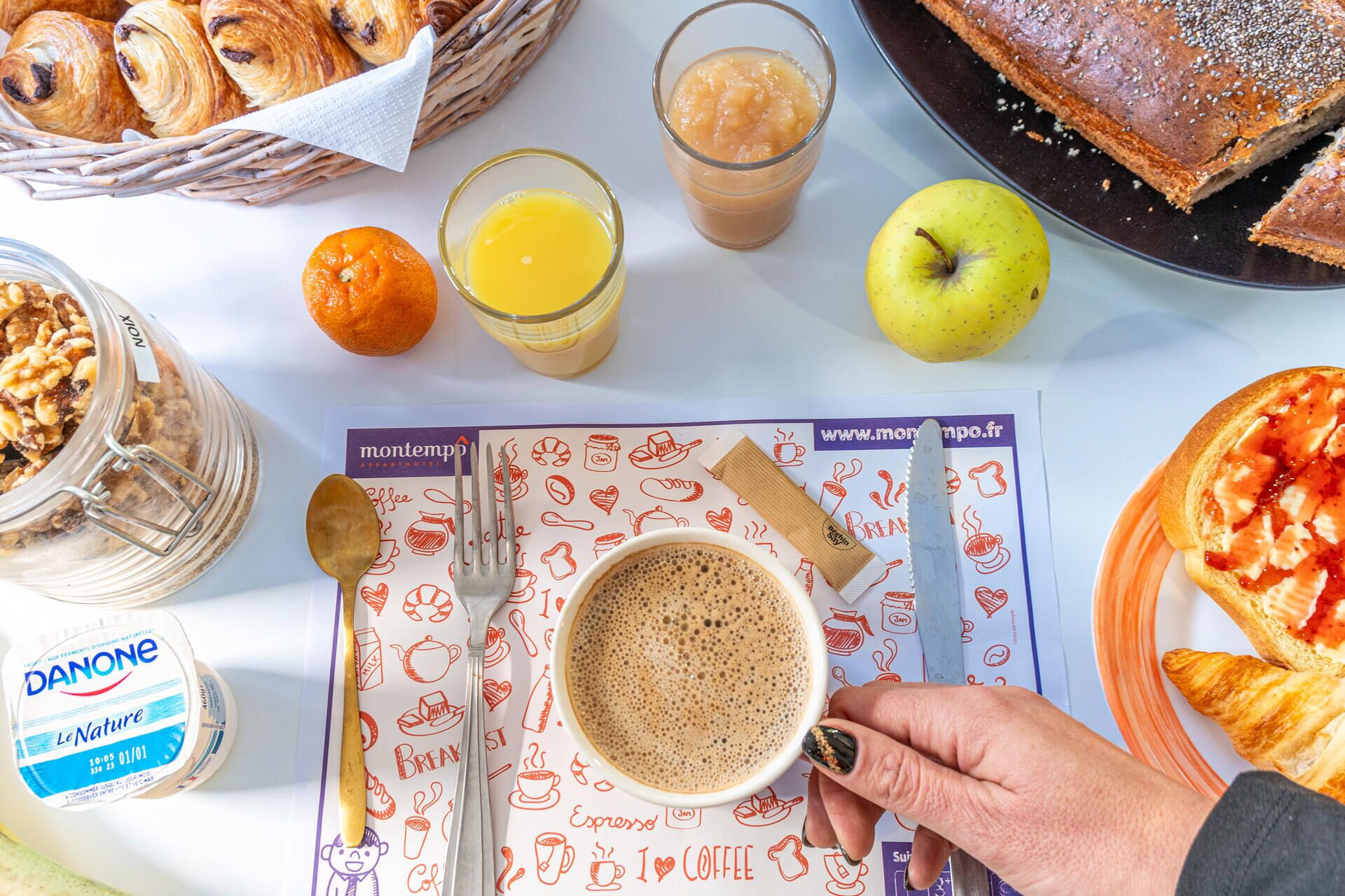 Mesa de café da manhã com croissants, suco de laranja, café e um pote de iogurte Danone.