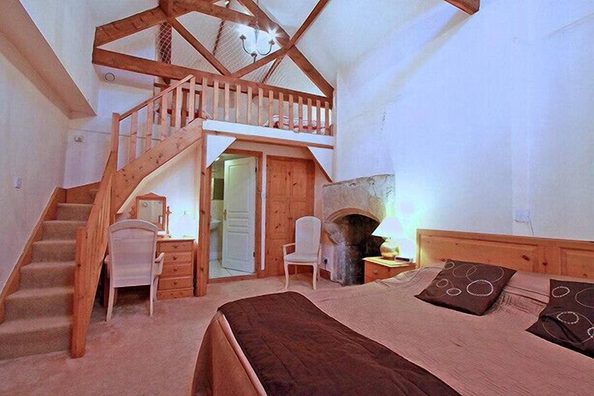 A bedroom with a wooden staircase, a fireplace, a bed, and a desk with a chair.