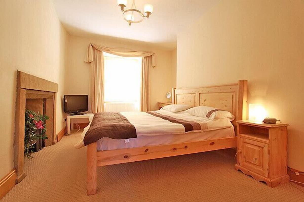 A bedroom with a wooden bed, a television on a stand, a nightstand, and a bedside table with a lamp.
