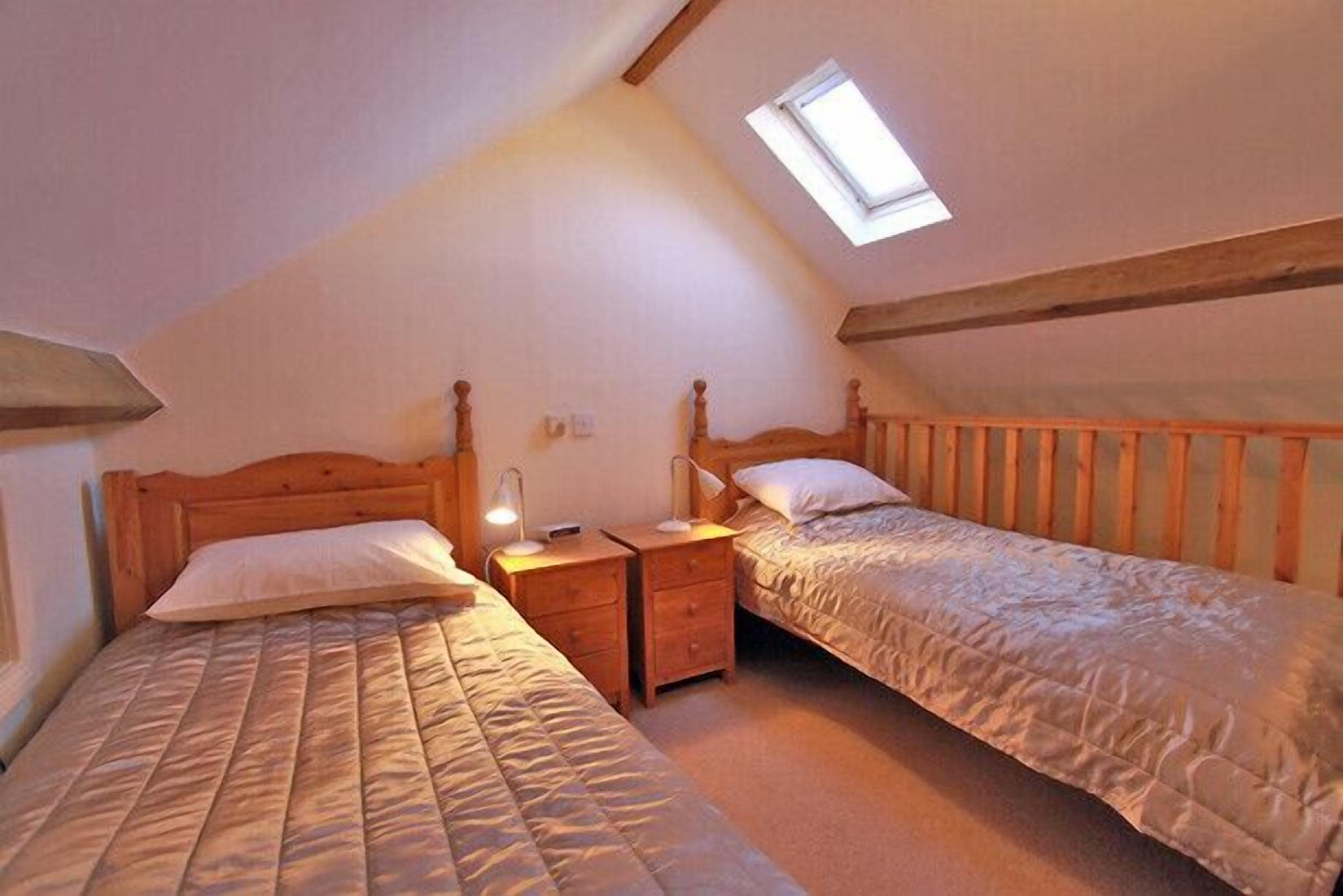 Een slaapkamer met twee bedden, houten hoofdeinden, een dakvenster en een klein nachtkastje met een lamp.