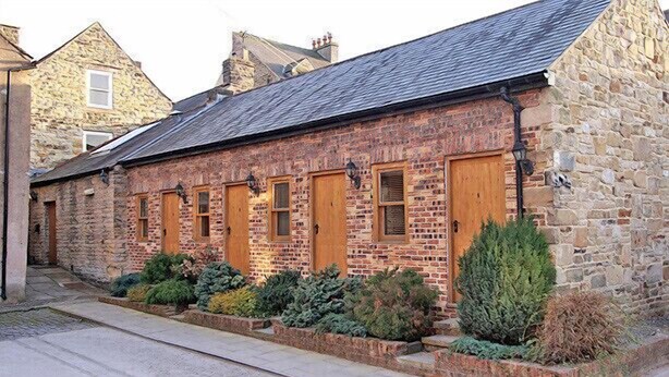 A stone building with a sloped roof, multiple wooden doors, and a small garden area with various plants. - Centre of Britain Hotel & Restaurant (Haltwhistle)