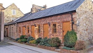 A stone building with a sloped roof, multiple wooden doors, and a small garden area with various plants.