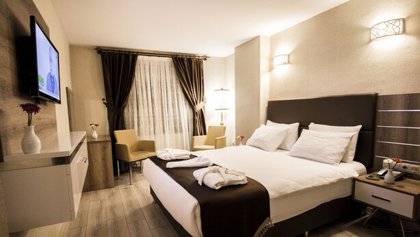 A modern hotel room with a large bed, bedside tables, a TV, and a seating area with a chair and a small table. - Hotel Business Han (Nevsehir)