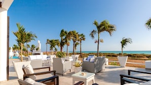 Un salón junto a la playa con sofás y mesas blancos, palmeras y vistas al océano.