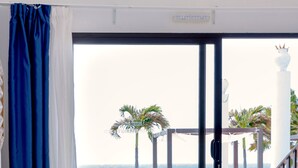 Habitación de hotel con cama, mesita de noche con lámpara, vista a la playa y balcón con mobiliario exterior.