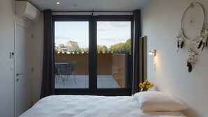 Une chambre avec un lit, une vue sur la ville grâce à de grandes fenêtres, un petit balcon avec une table et des chaises, et un attrape-rêves fixé au mur.