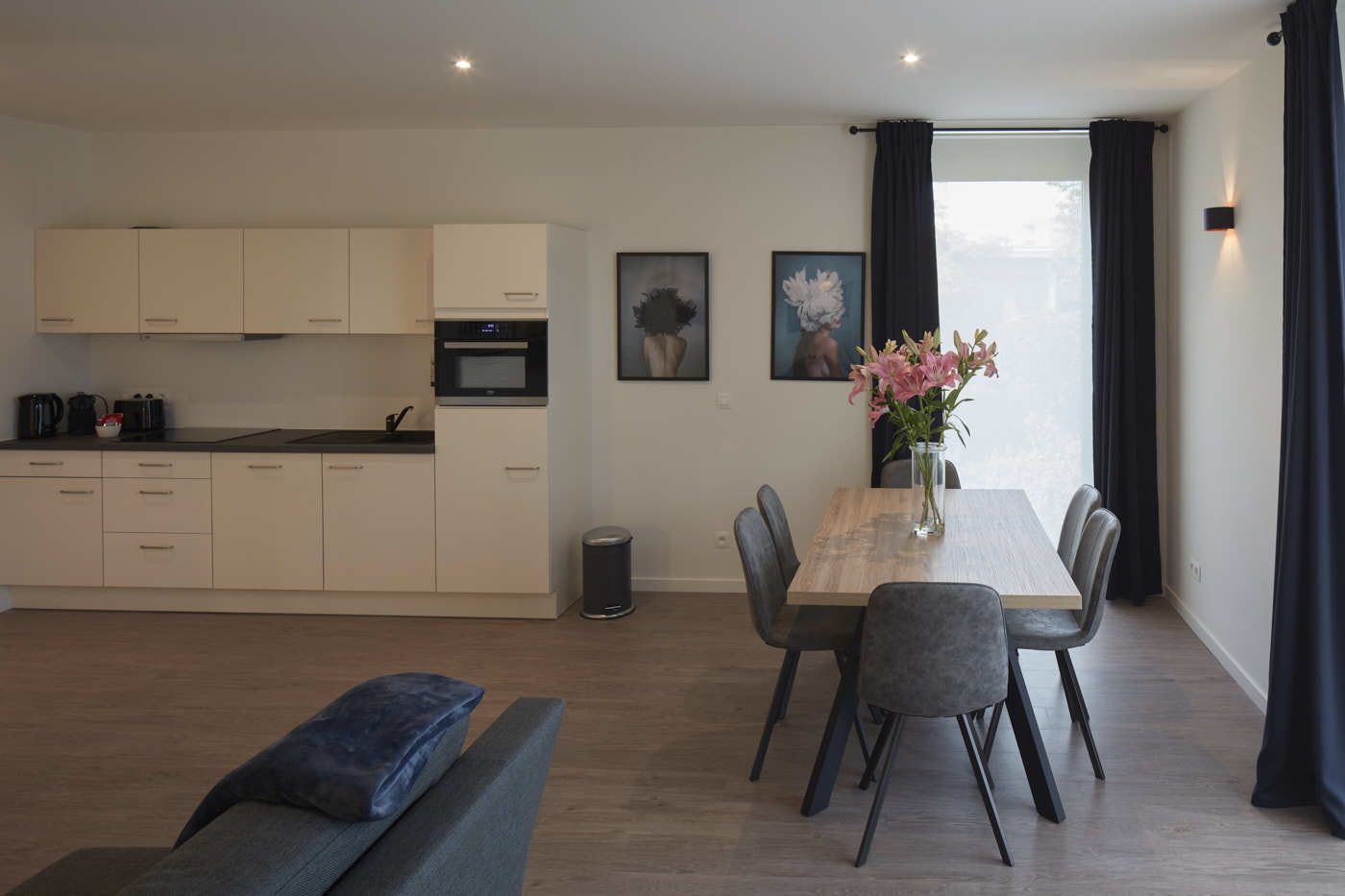 A modern kitchen with white cabinets and a built-in oven, a dining area with a wooden table and chairs, and a large window with curtains.