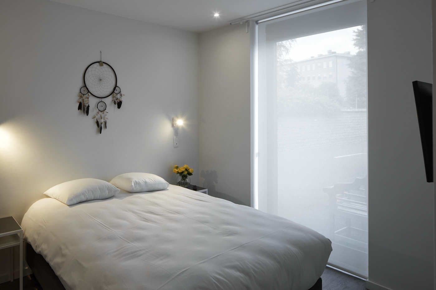 A bedroom with a large bed, a nightstand with a lamp, a dreamcatcher on the wall, and a window with a view of buildings.
